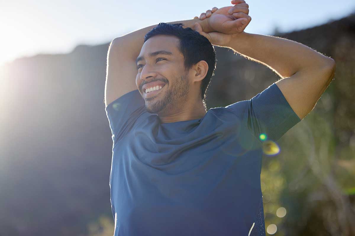 Homem sorrindo ao ar livre durante alongamento matinal, simbolizando pausa para autocuidado e equilíbrio emocional